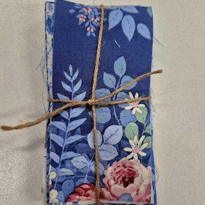 May include: A stack of rectangular fabric strips tied together with thin brown twine. The fabric features a vibrant blue background with a repeating pattern of light blue leaves, green foliage, and pink floral blossoms with white petals.
