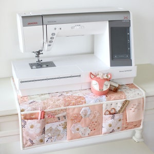 May include: A white Janome Horizon sewing machine with a grey top, positioned above a patchwork fabric organiser. The organiser features pockets containing sewing supplies, such as scissors and pens. A felt fox decoration is also present.