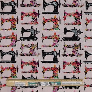 May include: Fabric with a repeating pattern of black sewing machines adorned with colorful floral arrangements. The sewing machines are set against a light pink background. A ruler is at the bottom of the image.