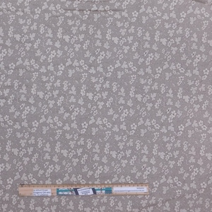 Könnte beinhalten: Beigefarbener Stoff mit einem sich wiederholenden Muster aus kleinen weißen Blumen und Blättern. Der Stoff hat eine subtile Textur und eine neutrale Farbpalette, geeignet für verschiedene Nähprojekte. Ein Lineal ist unten im Bild.