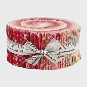 May include: A roll of fabric strips in shades of red and pink, tied with a white ribbon. The fabric features floral and geometric patterns. A measuring tape is wrapped around the roll, with the text "MODA Fabrics + Supplies".