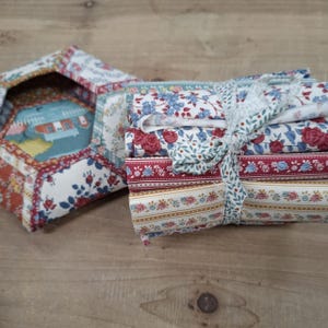 May include: A bundle of tightly rolled fabric strips featuring various floral and striped patterns in shades of red, blue, white, and mustard yellow. The collection is secured together with a matching fabric tie wrapped around the centre.