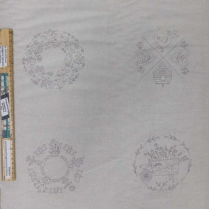May include: A light-coloured fabric featuring four embroidered garden-themed designs. The designs include a wreath of gardening tools, a bird with a birdhouse, a wreath of butterflies and flowers, and a wheelbarrow with flowers. A ruler is on the left side.