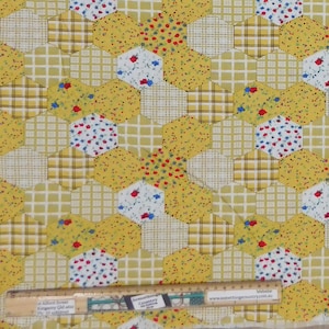 May include: Yellow fabric with a patchwork design featuring hexagons with a floral pattern, chequered pattern, and solid yellow. The fabric is measured with a ruler.