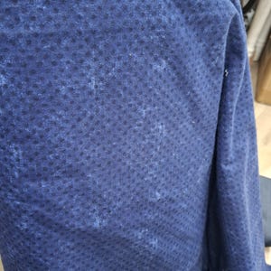 May include: Close-up of a navy blue fabric with a textured pattern of small, dark blue dots. The fabric has a slightly distressed appearance with lighter blue areas, creating a mottled effect. The material appears to be soft and possibly used for clothing.