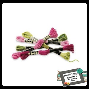 May include: Assortment of embroidery floss in pink, green, and white shades. The floss is wound on spools with black labels and gold lettering. The floss is arranged in a criss-cross pattern.