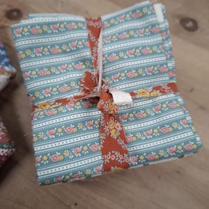 May include: A stack of folded fabric squares featuring various floral patterns in shades of blue, pink, cream, and rust. The fabric bundle is tied together with a strip of rust-coloured floral fabric, creating a neat, organised collection of textiles.