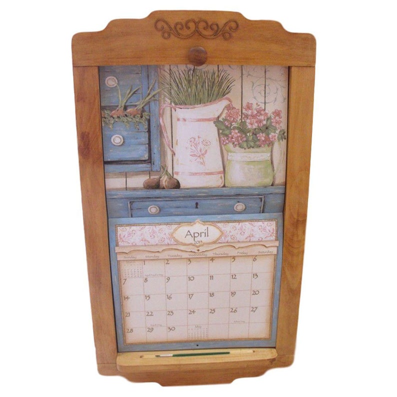 Puede incluir: Un calendario de madera con un dise&ntilde;o floral y un calendario para el mes de abril. El calendario est&aacute; unido a un marco de madera con un peque&ntilde;o estante en la parte inferior.