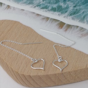 May include: A pair of silver heart shaped dangle earrings with a delicate chain. The earrings are on a wooden surface with a blue and white resin background.