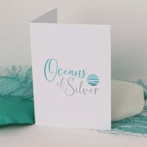 Pode incluir: Um cart&atilde;o branco com o texto "Oceans of Silver" em escrita turquesa e prateada, acompanhado por um logotipo circular de uma cena oce&acirc;nica estilizada. O cart&atilde;o est&aacute; parcialmente aberto, com tecido e rede turquesa no fundo.
