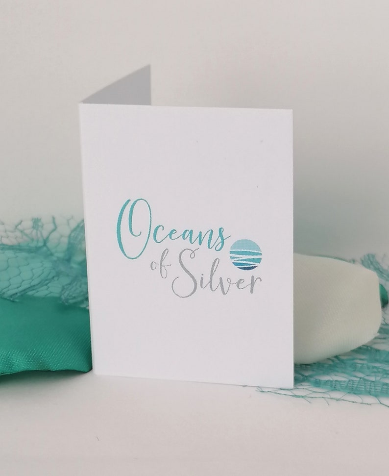 Pode incluir: Um cart&atilde;o branco com o texto "Oceans of Silver" em escrita turquesa e prateada, acompanhado por um logotipo circular de uma cena oce&acirc;nica estilizada. O cart&atilde;o est&aacute; parcialmente aberto, com tecido e rede turquesa no fundo.