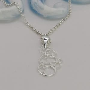 Peut inclure: Un collier en argent avec un pendentif représentant un motif abstrait de cercles interconnectés. La chaîne est délicate et le pendentif est en argent brillant. Le collier est présenté sur un fond clair.