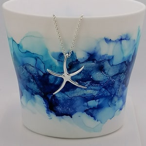 Peut inclure: Une tasse en c&eacute;ramique blanche avec un motif abstrait bleu et turquoise. Un collier en forme d'&eacute;toile de mer argent&eacute;e pend au bord de la tasse.