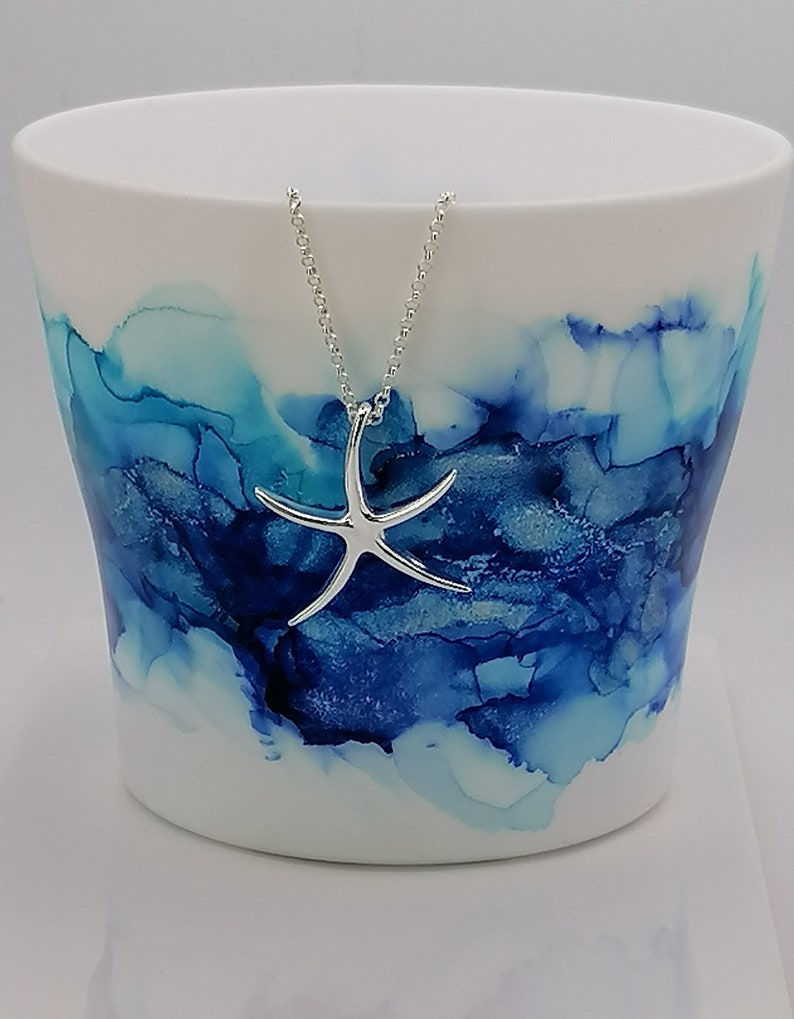 Peut inclure: Une tasse en c&eacute;ramique blanche avec un motif abstrait bleu et turquoise. Un collier en forme d'&eacute;toile de mer argent&eacute;e pend au bord de la tasse.