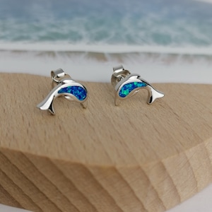 May include: A pair of silver dolphin stud earrings with blue opal accents. The earrings are on a wooden surface.