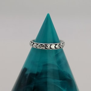 May include: A silver ring featuring a repeating heart design. The ring is displayed on a teal-colored, cone-shaped stand against a neutral background. The ring's design consists of small, evenly spaced hearts around the band.