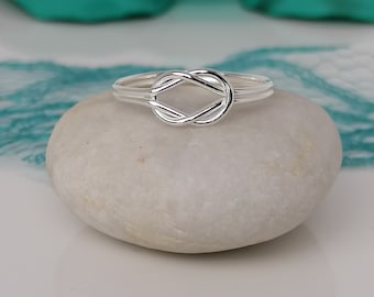 May include: A silver knot ring with a simple design. The ring is on a white rock.