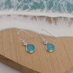 Peut inclure: Boucles d'oreilles pendantes argentées avec des pierres bleues claires en forme de goutte d'eau. Les boucles d'oreilles sont présentées sur une surface en bois, avec un arrière-plan flou de vagues bleues et blanches.