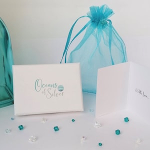 Pode incluir: Uma caixa de presente branca com o texto "Oceans of Silver" em escrita turquesa, uma sacola de presente de organza turquesa e um cart&atilde;o branco com "With Love..." escrito dentro. Pequenos cristais transparentes e turquesas est&atilde;o espalhados.