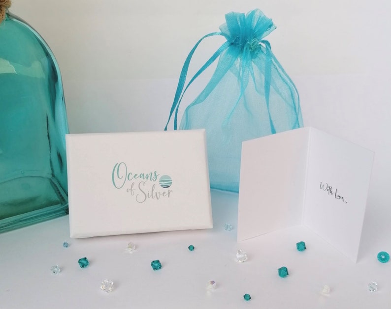 Pode incluir: Uma caixa de presente branca com o texto "Oceans of Silver" em escrita turquesa, uma sacola de presente de organza turquesa e um cart&atilde;o branco com "With Love..." escrito dentro. Pequenos cristais transparentes e turquesas est&atilde;o espalhados.