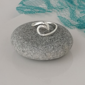 Pode incluir: Um anel de prata em forma de onda sobre uma pedra cinza. O anel é um design simples com forma de onda.