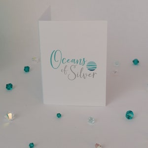 Peut inclure: Une carte blanche avec le texte "Oceans of Silver" en bleu et gris. Le texte est en &eacute;criture cursive et est centr&eacute; sur la carte. Il y a un petit cercle bleu et gris avec un motif de vague &agrave; droite du texte.
