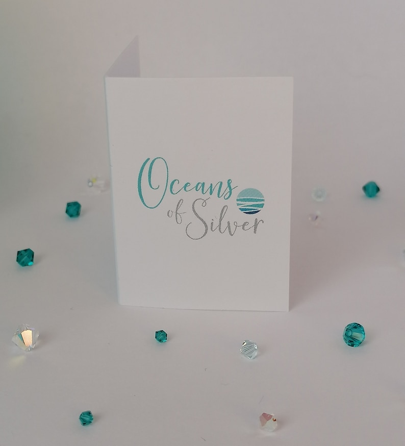 Peut inclure: Une carte blanche avec le texte "Oceans of Silver" en bleu et gris. Le texte est en &eacute;criture cursive et est centr&eacute; sur la carte. Il y a un petit cercle bleu et gris avec un motif de vague &agrave; droite du texte.