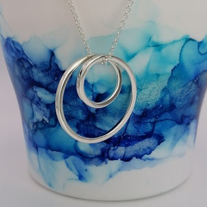 May include: A silver pendant necklace featuring two intertwined, open circles. The pendant hangs from a delicate chain. The background is a white ceramic cup with blue and white abstract ink patterns.