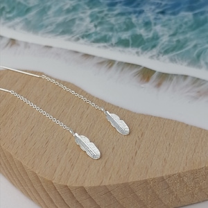 Puede incluir: Un par de pendientes de plumas de plata con cadenas delicadas. Los pendientes se muestran sobre una superficie de madera con un fondo de resina azul y blanco.