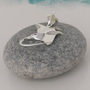 Pode incluir: Um pingente de prata polida em forma de arraia manta, repousando sobre uma pedra cinza. O pingente tem um design detalhado, com as asas e o corpo da arraia claramente definidos. O fundo &eacute; uma mistura suave e desfocada de branco e turquesa.