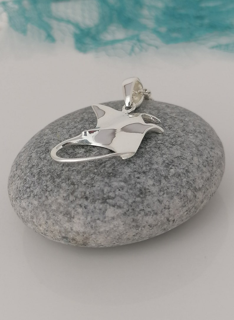 Pode incluir: Um pingente de prata polida em forma de arraia manta, repousando sobre uma pedra cinza. O pingente tem um design detalhado, com as asas e o corpo da arraia claramente definidos. O fundo &eacute; uma mistura suave e desfocada de branco e turquesa.