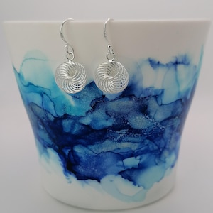 Peut inclure: Paire de boucles d'oreilles pendantes argentées en spirale. Chaque boucle d'oreille est suspendue à un crochet et présente un motif sphérique avec un motif ajouré détaillé. Les boucles d'oreilles sont présentées sur un fond abstrait blanc et bleu.