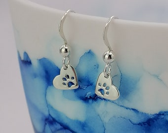 May include: A pair of silver dangle earrings with a heart-shaped charm featuring a paw print cutout. The earrings are hanging from a white ceramic mug with blue and white marbling.