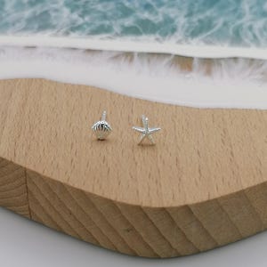 May include: Two small silver earrings, one shaped like a seashell and the other like a starfish, sit on a wooden surface. The background features a blurred image of a beach scene with blue water and white foam. These earrings are perfect for beach lovers.