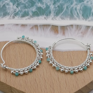 Puede incluir: Pendientes de aro de plata con un diseño delicado que presenta pequeñas cuentas turquesas. Los pendientes están sobre una superficie de madera.