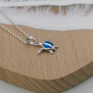 Könnte beinhalten: Eine silberne Halskette mit einem blauen Opal-Schildkröten-Anhänger. Die Halskette liegt auf einer Holzoberfläche mit weißem Hintergrund.