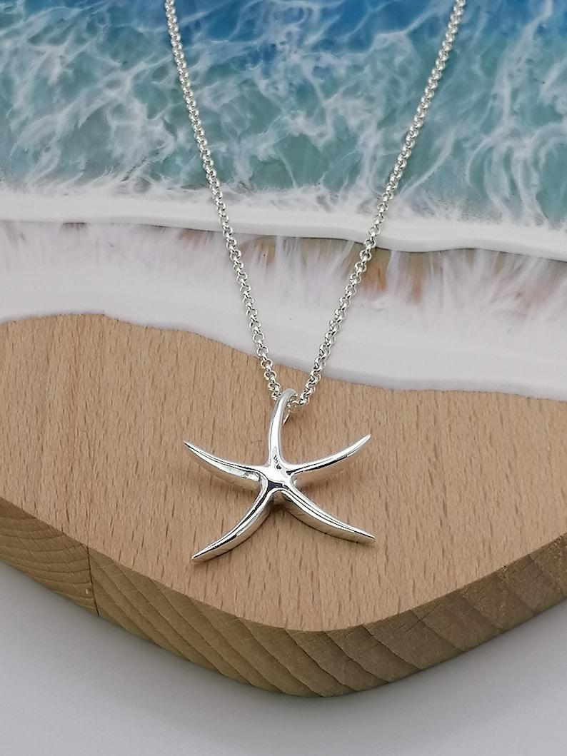 Peut inclure: Un collier en argent avec un pendentif en forme d'&eacute;toile de mer. Le collier est pos&eacute; sur une surface en bois avec un fond bleu et blanc.