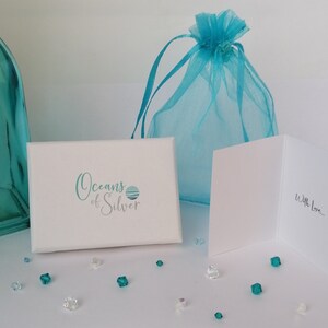 Peut inclure: Une bo&icirc;te cadeau blanche avec le texte "Oceans of Silver" et un motif de vague bleu et blanc. La bo&icirc;te est entour&eacute;e de petits cristaux bleus et transparents. Un sac en organza bleu et une carte blanche avec le texte "With Love..." sont &eacute;galement visibles.
