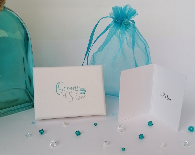 Peut inclure: Une bo&icirc;te cadeau blanche avec le texte "Oceans of Silver" et un motif de vague bleu et blanc. La bo&icirc;te est entour&eacute;e de petits cristaux bleus et transparents. Un sac en organza bleu et une carte blanche avec le texte "With Love..." sont &eacute;galement visibles.