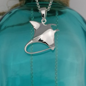 Pode incluir: Pingente de prata em forma de arraia manta, pendurado em uma corrente fina. O pingente polido reflete a luz, destacando-se em um fundo turquesa desfocado. A corrente tem um design delicado, real&ccedil;ando a eleg&acirc;ncia da pe&ccedil;a.