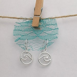 May include: Silver earrings with a wave design hanging from a turquoise mesh fabric. The earrings are hanging from a wooden clothespin.
