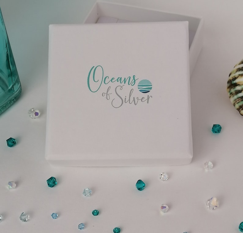 Pode incluir: Uma caixa de joias branca com o texto "Oceans of Silver" em escrita turquesa e um logotipo turquesa. Pequenos cristais iridescentes e turquesas est&atilde;o espalhados ao redor da caixa. Uma garrafa de vidro turquesa &eacute; vis&iacute;vel.