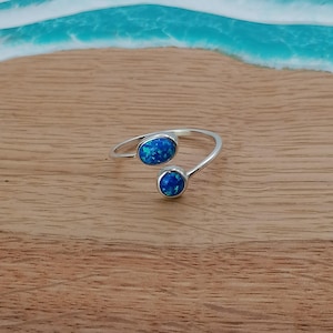 May include: A silver ring with two blue opal stones, one larger than the other. The ring has an open design. The ring is displayed on a wooden surface, with a blurred background of blue and white, reminiscent of waves.