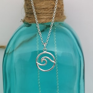 May include: A silver necklace with a wave pendant. The pendant is a circle with a wave design inside. The necklace is hanging on a blue glass bottle.