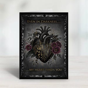 Op de afbeelding: Een wenskaart met een zwart hartontwerp, versierd met gouden aderen en donkerrode rozen. De kaart heeft de tekst "EVEN IN DARKNESS.....MY HEART FINDS YOU" in gouden letters, tegen een donkergrijze achtergrond met een decoratieve rand.