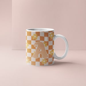 Puede incluir: Taza de cerámica blanca con un patrón a cuadros en naranja y blanco. La taza tiene una gran letra "A" naranja estilizada en el centro y pequeñas flores amarillas sobre el fondo a cuadros.