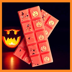 May include: Two orange wax melts shaped like a bar with a pumpkin design. Each bar is divided into nine squares with a pumpkin face in each square. The wax melts are on a dark background.