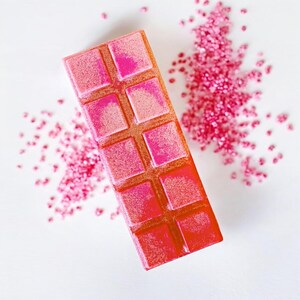May include: A pink and orange wax melt bar with a grid pattern. The bar is surrounded by pink glitter.