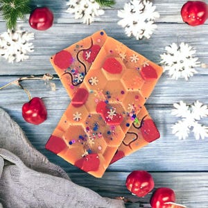 May include: Two wax melts in a honeycomb pattern with red and white snowflake designs. The wax melts are on a light blue wooden surface with red cherries and white snowflakes.