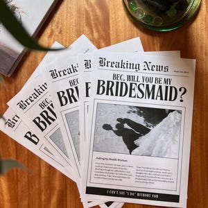 May include: A mock newspaper announcement with the headline "Breaking News" and the text "BEC, WILL YOU BE MY BRIDESMAID?"  The announcement is printed on white paper with black text. The announcement is folded in half and is sitting on a wooden table.